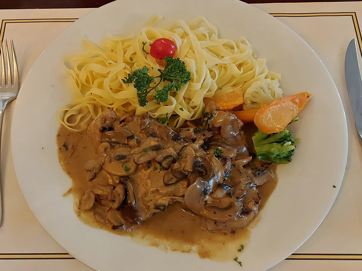 Two pork cutlets with fresh mushroom cream sauce, served with tagliatelle, accompanied by cauliflower, carrots, and broccoli, garnished with parsley and a half cherry tomato.