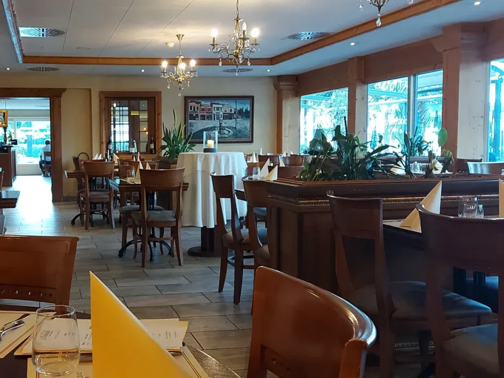 Interior of Ambiente Bern: A bright room with many windows, dark wooden tables set with high-quality paper napkins, and matching dark wooden chairs with seat cushions. The walls are decorated with pictures of Italian motifs.