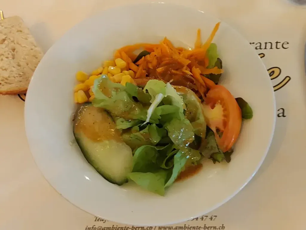 Small lunch salad on a white deep plate, with green lettuce leaves, a slice of tomato, carrots, corn, and a piece of cucumber, dressed with Italian salad dressing.