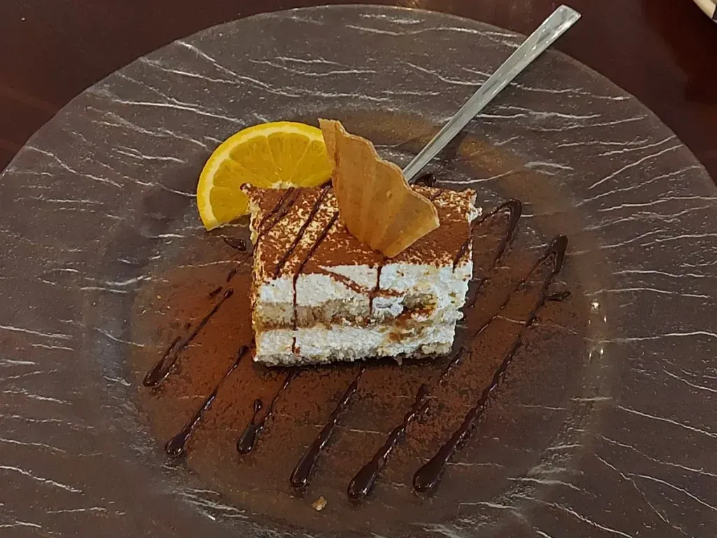 A cuboid piece of tiramisu, garnished with a cookie wafer, served on a round, textured glass plate, decorated with cocoa powder, chocolate sauce stripes, and a half orange slice.