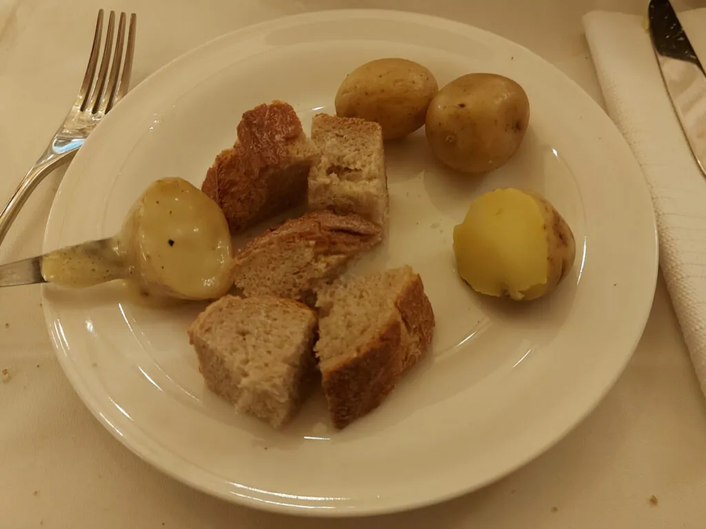 Nahaufnahme eines Tellers mit Brotwürfeln, Gschwellti und einer Fonduegabel, an der eine mit flüssigem Käse überzogene Kartoffel steckt