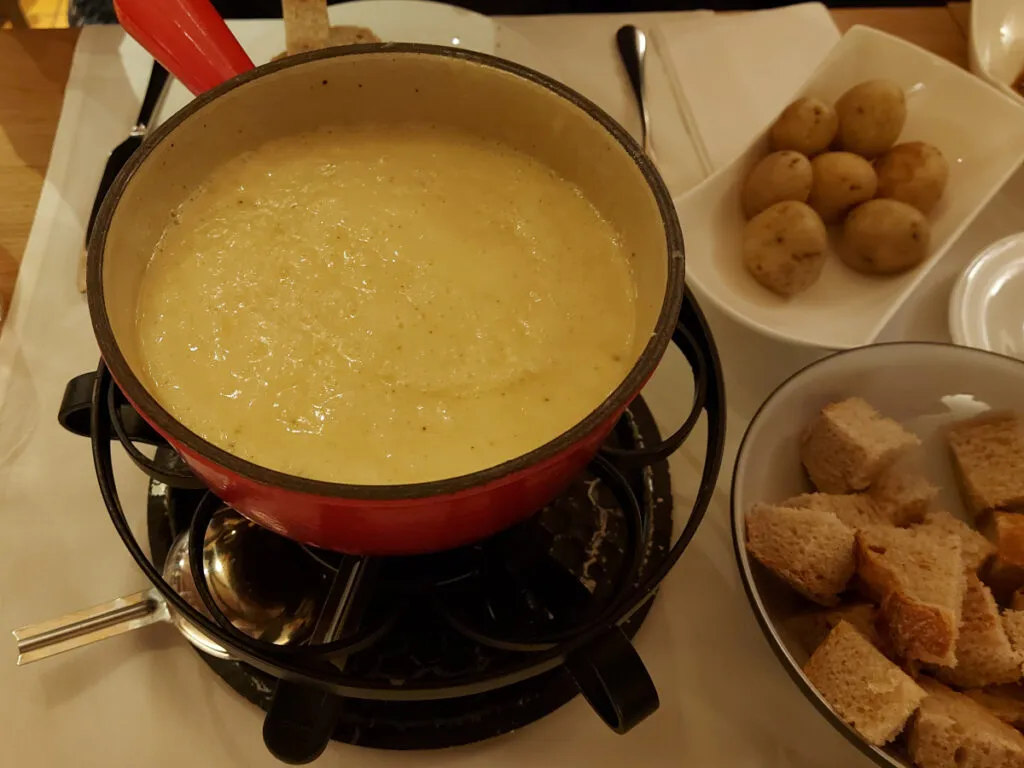 Sämiges Käsefondue im roten Caquelon auf einem Rechaud, serviert mit Brotwürfeln und kleinen Kartoffeln (Gschwellti)